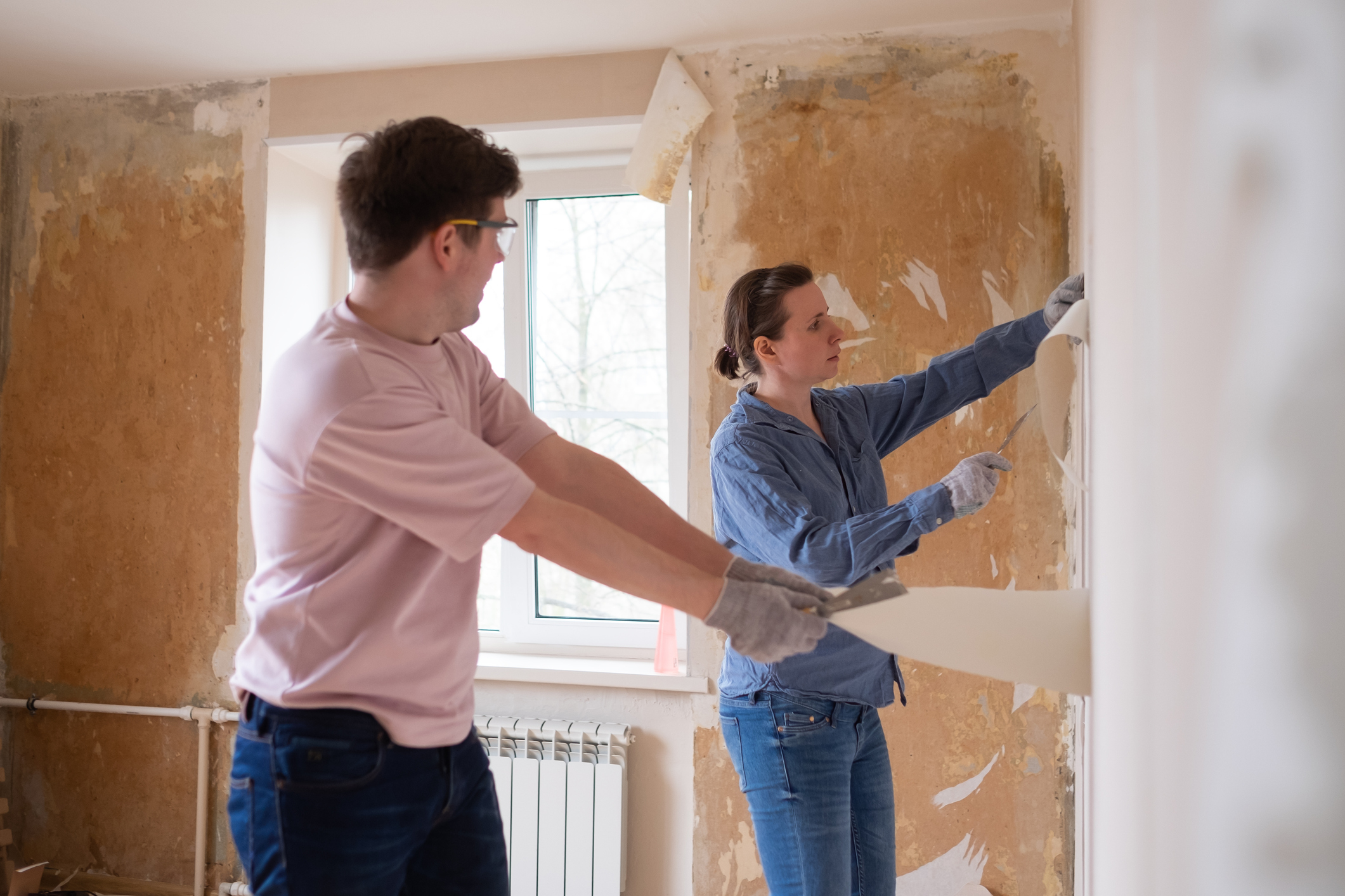 Young couple removing old wallpaper from walls preparing for flat renovation.